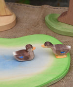 Two Ostheimer wooden ducks placed on a wooden pond scene piece. One duck is in a swimming position one is standing and has a long neck position.