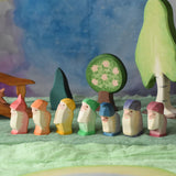 A collection of 7 coloured Ostheimer wooden dwraf wooden toy figures placed in a line in a small world play scene. There are dwarves each with a different coloured outfit, red, orange, yellow, green, blue, purple and violet. They all have matching coloured hats and white beards. There are wooden trees in the background including an apple tree and the large birch tree. These wooden figures are part of a large range of expertly hand crafted, Waldorf inspired, wooden toys here at Babipur. 