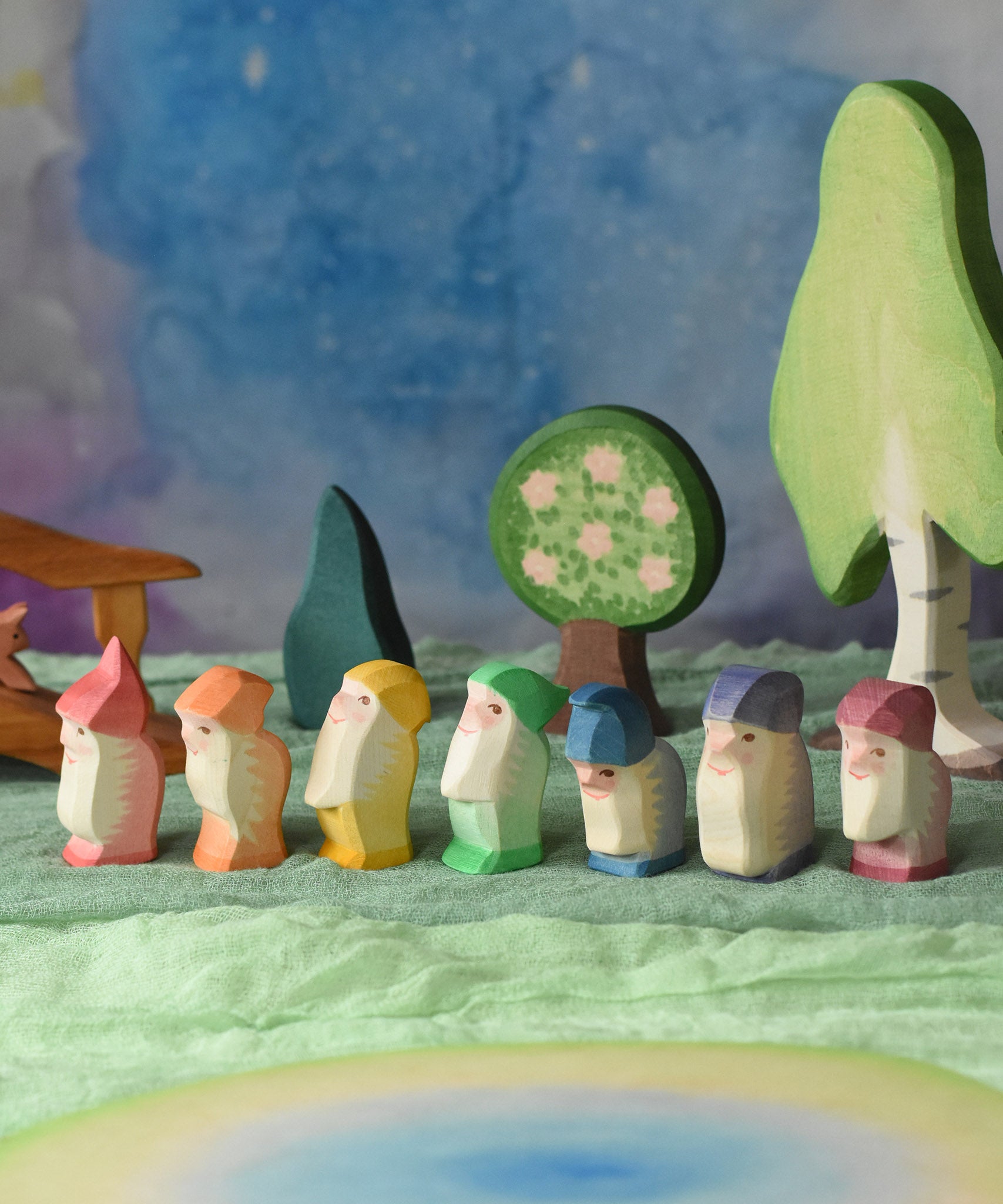 A collection of 7 coloured Ostheimer wooden dwraf wooden toy figures placed in a line in a small world play scene. There are dwarves each with a different coloured outfit, red, orange, yellow, green, blue, purple and violet. They all have matching coloured hats and white beards. There are wooden trees in the background including an apple tree and the large birch tree. These wooden figures are part of a large range of expertly hand crafted, Waldorf inspired, wooden toys here at Babipur. 