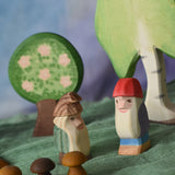 An Ostheimer fire tree dwarf placed next to a Bodo dwarf in a small world play scene. Ostheimer wooden trees can be seen in the background with a green Ostheimer scarf covering the surface that they are placed upon. There are three Grapat wooden mushroom mandala pieces at the front of the scene. 
These wooden figures are part of a large range of expertly hand crafted, Waldorf inspired, wooden toys here at Babipur.