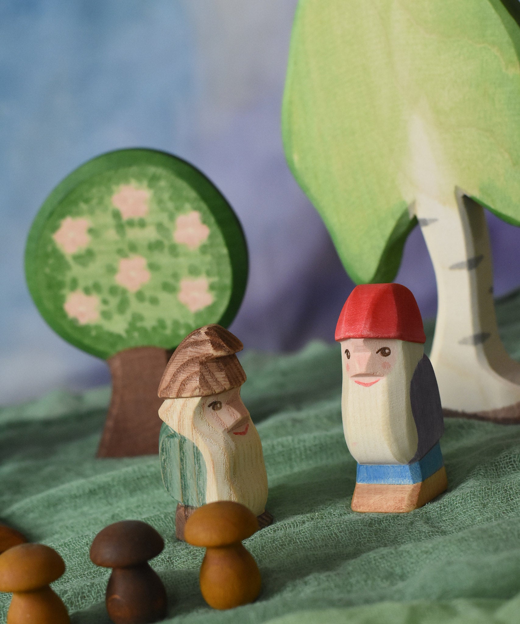 An Ostheimer fire tree dwarf placed next to a Bodo dwarf in a small world play scene. Ostheimer wooden trees can be seen in the background with a green Ostheimer scarf covering the surface that they are placed upon. There are three Grapat wooden mushroom mandala pieces at the front of the scene. 
These wooden figures are part of a large range of expertly hand crafted, Waldorf inspired, wooden toys here at Babipur.