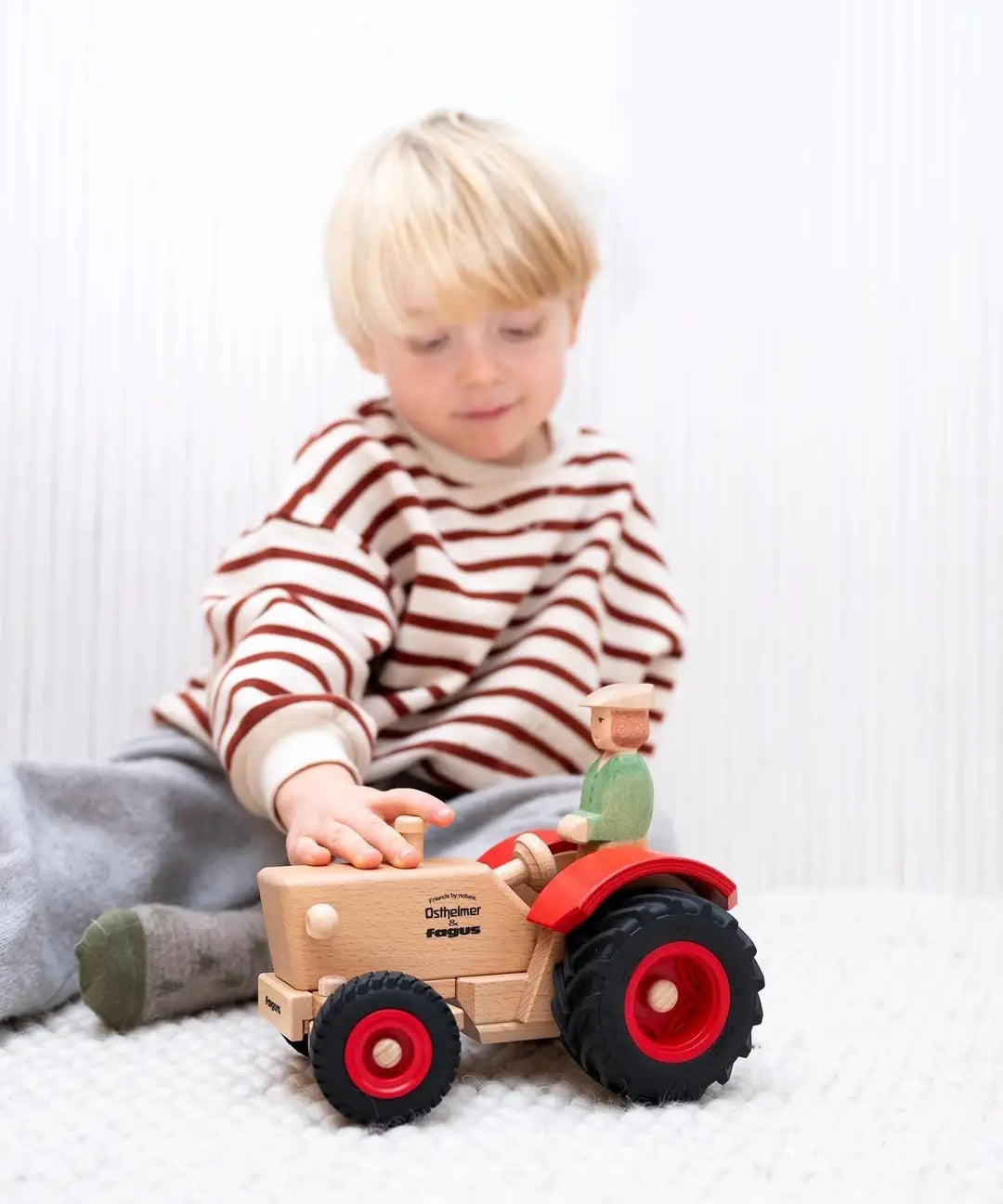 Child playing with the Ostheimer x Fagus wooden tractor and trailer indoors, with the farmer figure seated in the tractor.