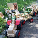 Ostheimer x Fagus wooden toy tractor pulling a Fagus trailer through an outdoor farm play scene with Ostheimer animals and wooden farm vehicles.