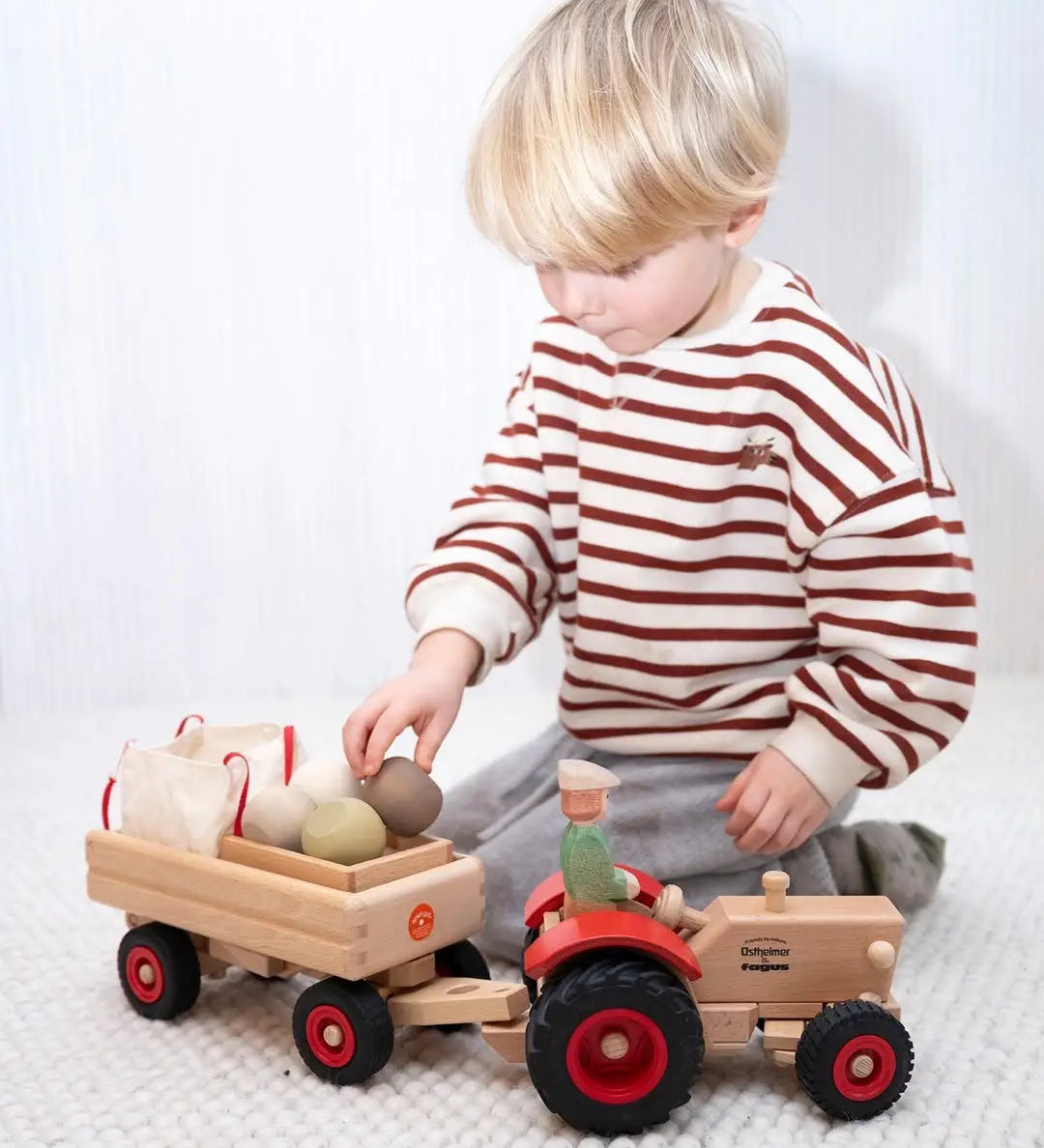 Child loading wooden sacks and vegetables into a Fagus trailer attached to the Ostheimer x Fagus toy tractor.