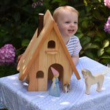A child standing behind an Ostheimer Fairy Tale wooden dolls house. A medieval princess, cat and horse figures are placed in front of the house. These wooden figures are part of a large range of expertly hand crafted, Waldorf inspired, wooden toys here at Babipur.