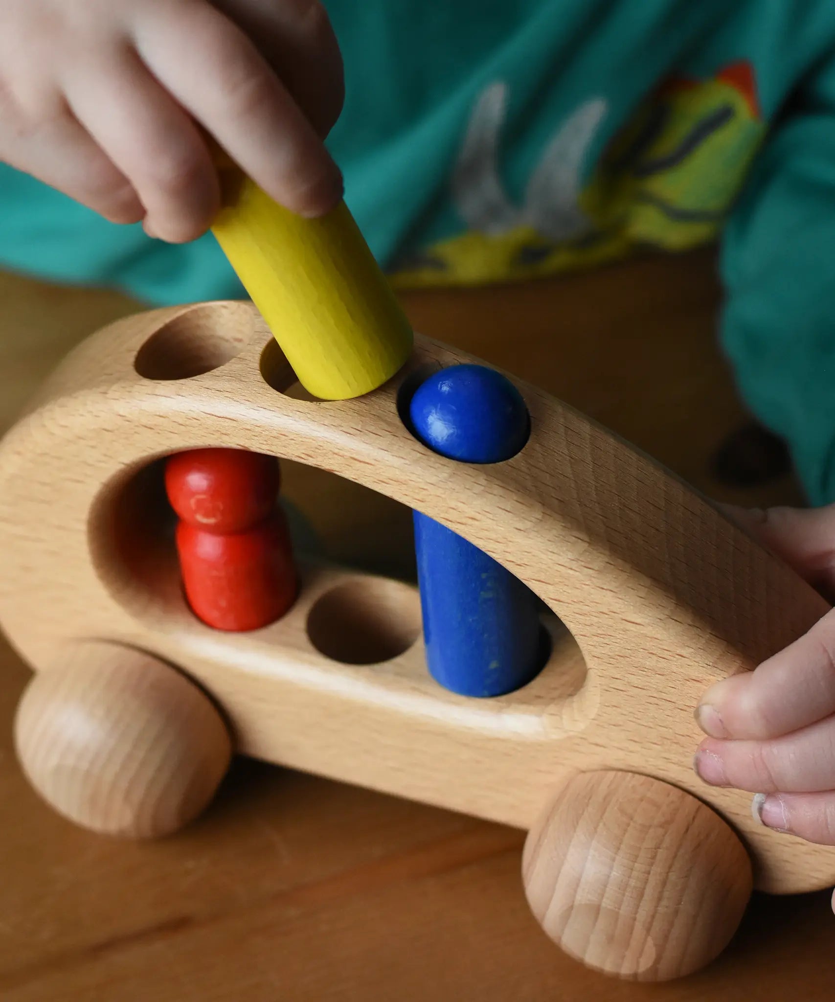 Close up of a child's hand placing the yellow peg doll into the circular slots in the Ostheimer family van push along wooden toy.