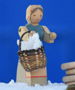 An Ostheimer Farmer Woman handcrafted wooden toy figure placed on a snow covered surface in the Babipur garden. The wooden toy figure is holding a small kraul basket filled with snow with it's pose-able arms. These wooden figures are part of a large range of expertly hand crafted, Waldorf inspired, wooden toys here at Babipur.