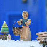 An Ostheimer Farmer Woman handcrafted wooden toy figure placed on a snow covered surface in the Babipur garden with a small kraul basket hung on it's arm and a Mader tree shaped spinning top on a branch placed next to them. These wooden figures are part of a large range of expertly hand crafted, Waldorf inspired, wooden toys here at Babipur.