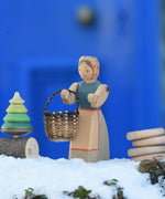 An Ostheimer Farmer Woman handcrafted wooden toy figure placed on a snow covered surface in the Babipur garden with a small kraul basket hung on it's arm and a Mader tree shaped spinning top on a branch placed next to them. These wooden figures are part of a large range of expertly hand crafted, Waldorf inspired, wooden toys here at Babipur.