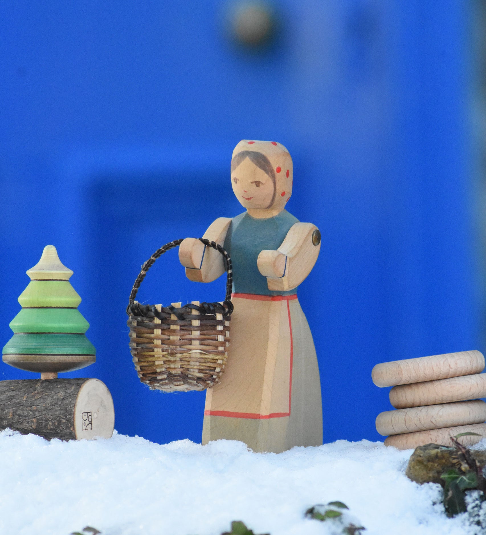 An Ostheimer Farmer Woman handcrafted wooden toy figure placed on a snow covered surface in the Babipur garden with a small kraul basket hung on it's arm and a Mader tree shaped spinning top on a branch placed next to them. These wooden figures are part of a large range of expertly hand crafted, Waldorf inspired, wooden toys here at Babipur.