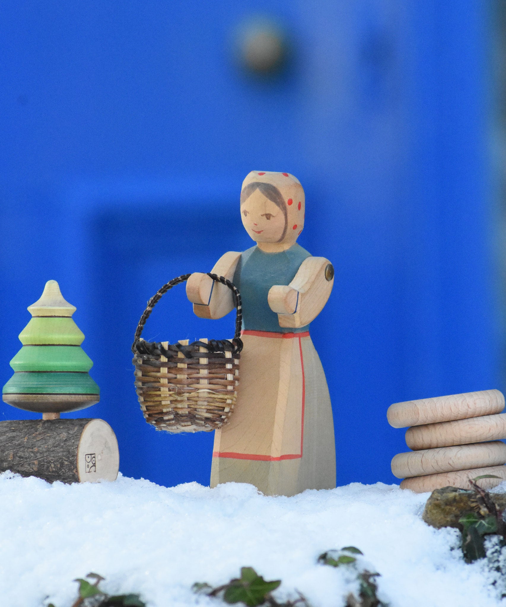 An Ostheimer Farmer Woman handcrafted wooden toy figure placed on a snow covered surface in the Babipur garden with a small kraul basket hung on it's arm and a Mader tree shaped spinning top on a branch placed next to them. These wooden figures are part of a large range of expertly hand crafted, Waldorf inspired, wooden toys here at Babipur.