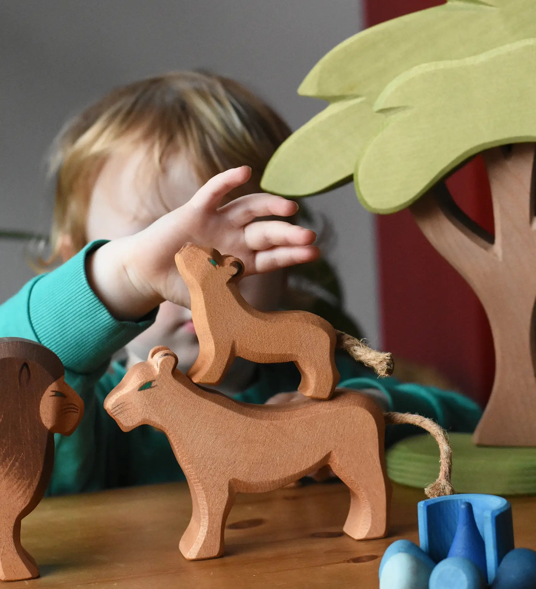 Ostheimer cub placed on a female lioness wooden toy figure with a child reaching for the cub figure in a small world play scene with the African tree.