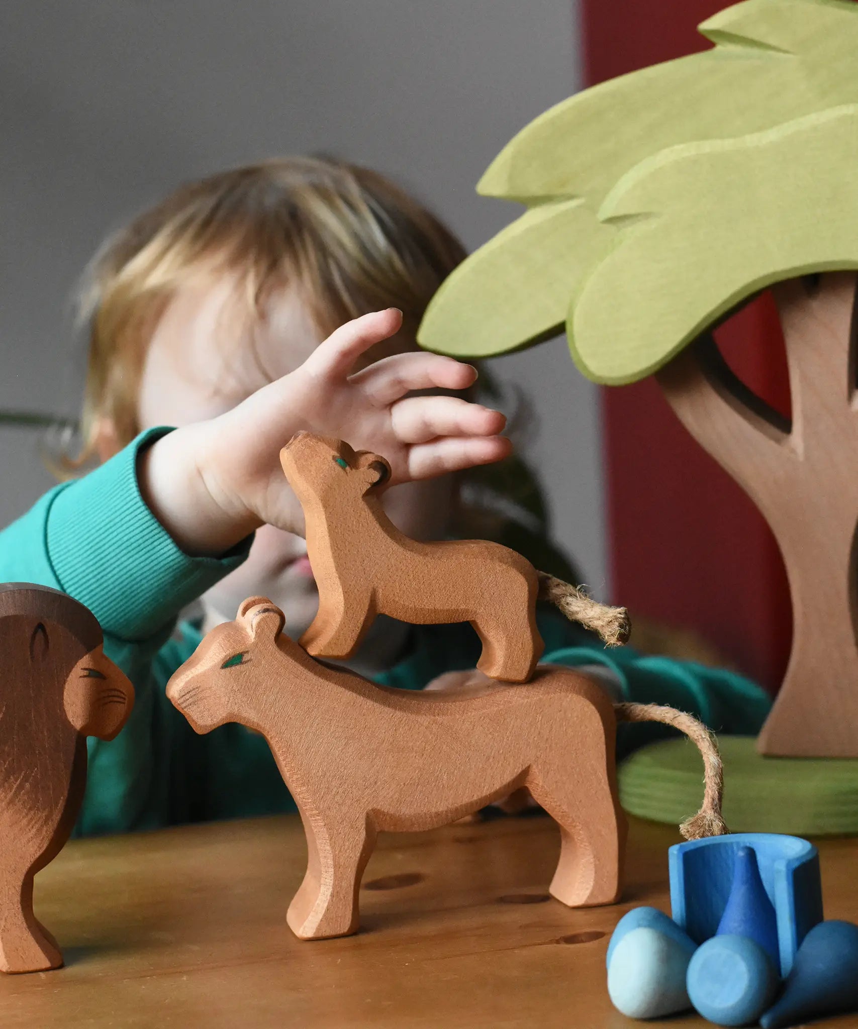 Ostheimer cub placed on a female lioness wooden toy figure with a child reaching for the cub figure in a small world play scene with the African tree.