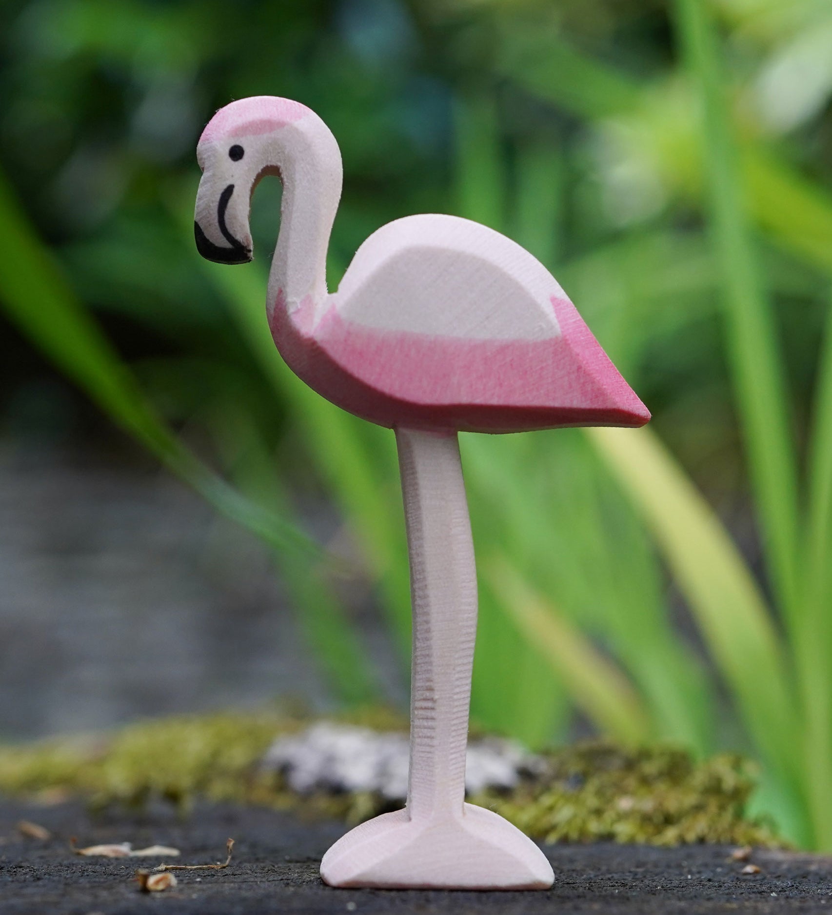 An Ostheimer flamingo bird placed on a mossy surface outdoors, blades of grass can be seen blurred in the background. This expertly hand crafted wooden figure is part of a large range of wooden toys here at Babipur.
