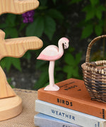 An Ostheimer flamingo wooden toy figure placed on a pile of books. An Otheimer bird tree can be seen in the background.