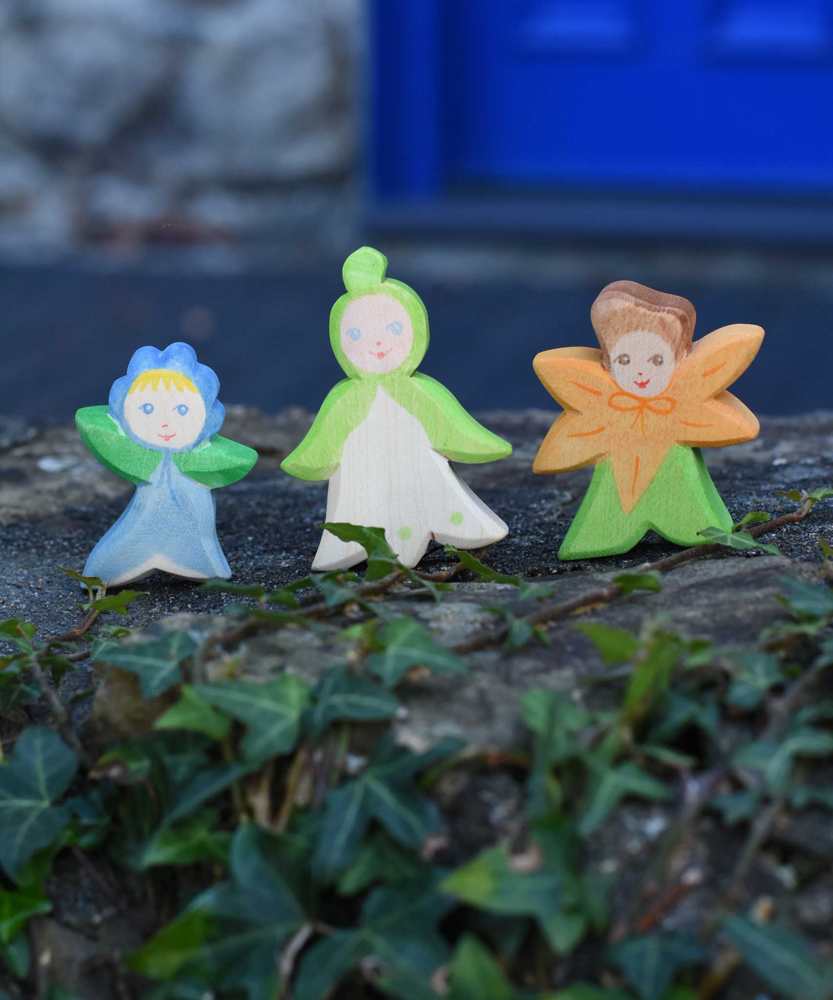 Three Ostheimer hand made and hand painted wooden forget-me-not, snowdrop and calendula Flower Child figure toys placed on a stone wall outdoors. These flower child wooden figures are part of a wide range of expertly hand crafted wooden toys here at Babipur. 