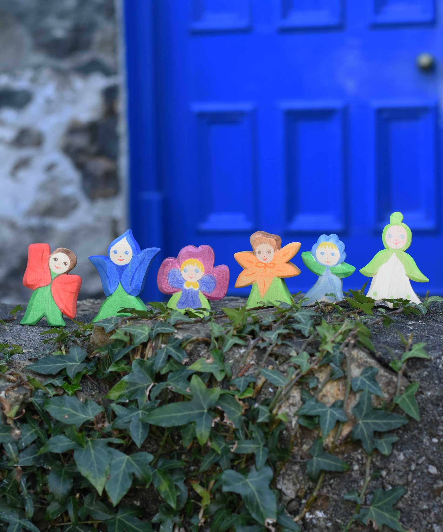 Six Ostheimer hand made and hand painted wooden poppy Flower Child figure toys placed on a stone wall outdoors. The six flower child figures include the gentian, forget-me-not, pansy, calendula, snowdrop and poppy. These flower child wooden figures are part of a wide range of expertly hand crafted wooden toys here at Babipur. 