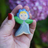 An Ostheimer hand made and hand painted wooden forget-me-not Flower Child figure toy held in an adult's hand to show scale. These flower child wooden figures are part of a wide range of expertly hand crafted wooden toys here at Babipur. 