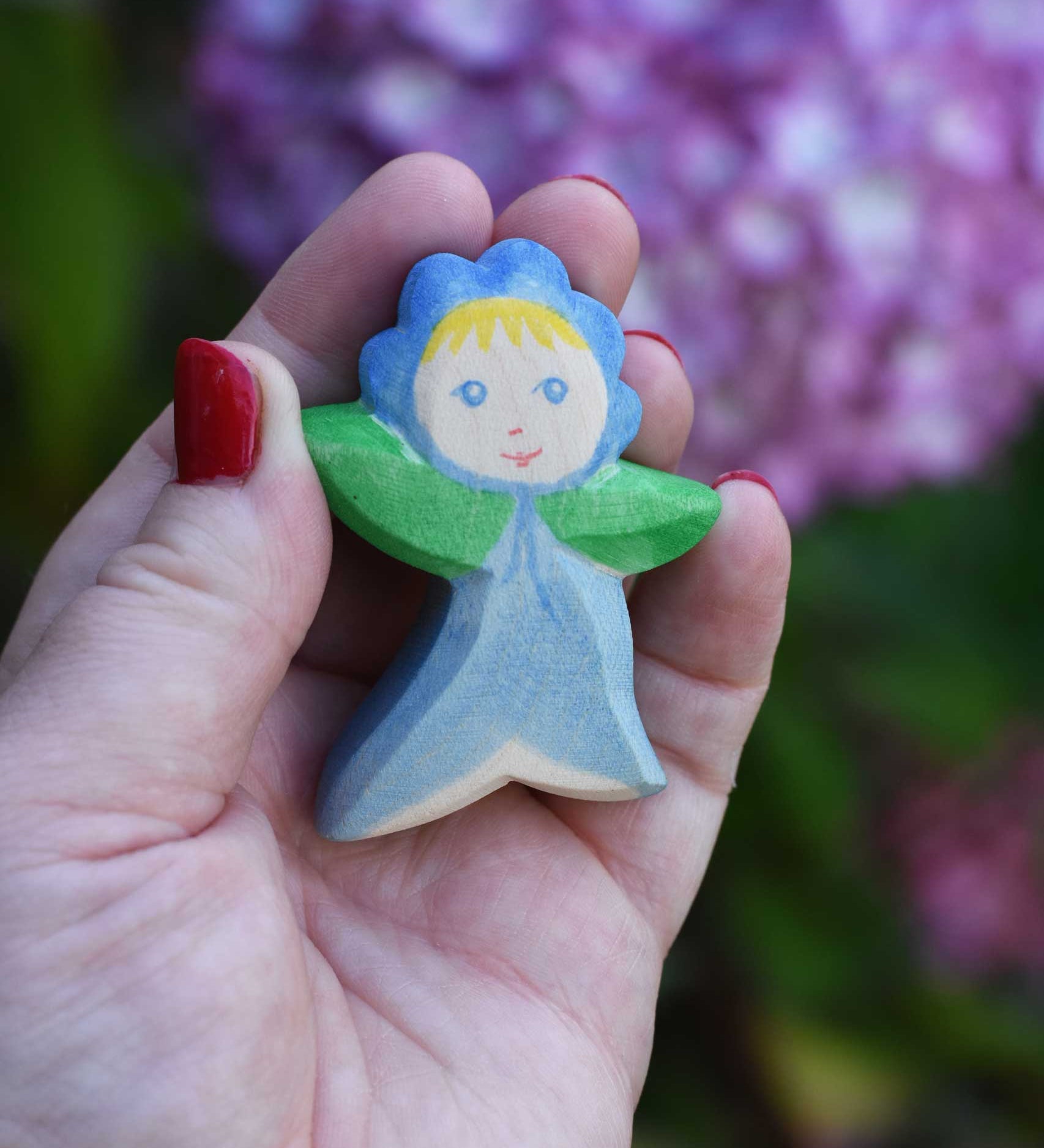 An Ostheimer hand made and hand painted wooden forget-me-not Flower Child figure toy held in an adult's hand to show scale. These flower child wooden figures are part of a wide range of expertly hand crafted wooden toys here at Babipur. 