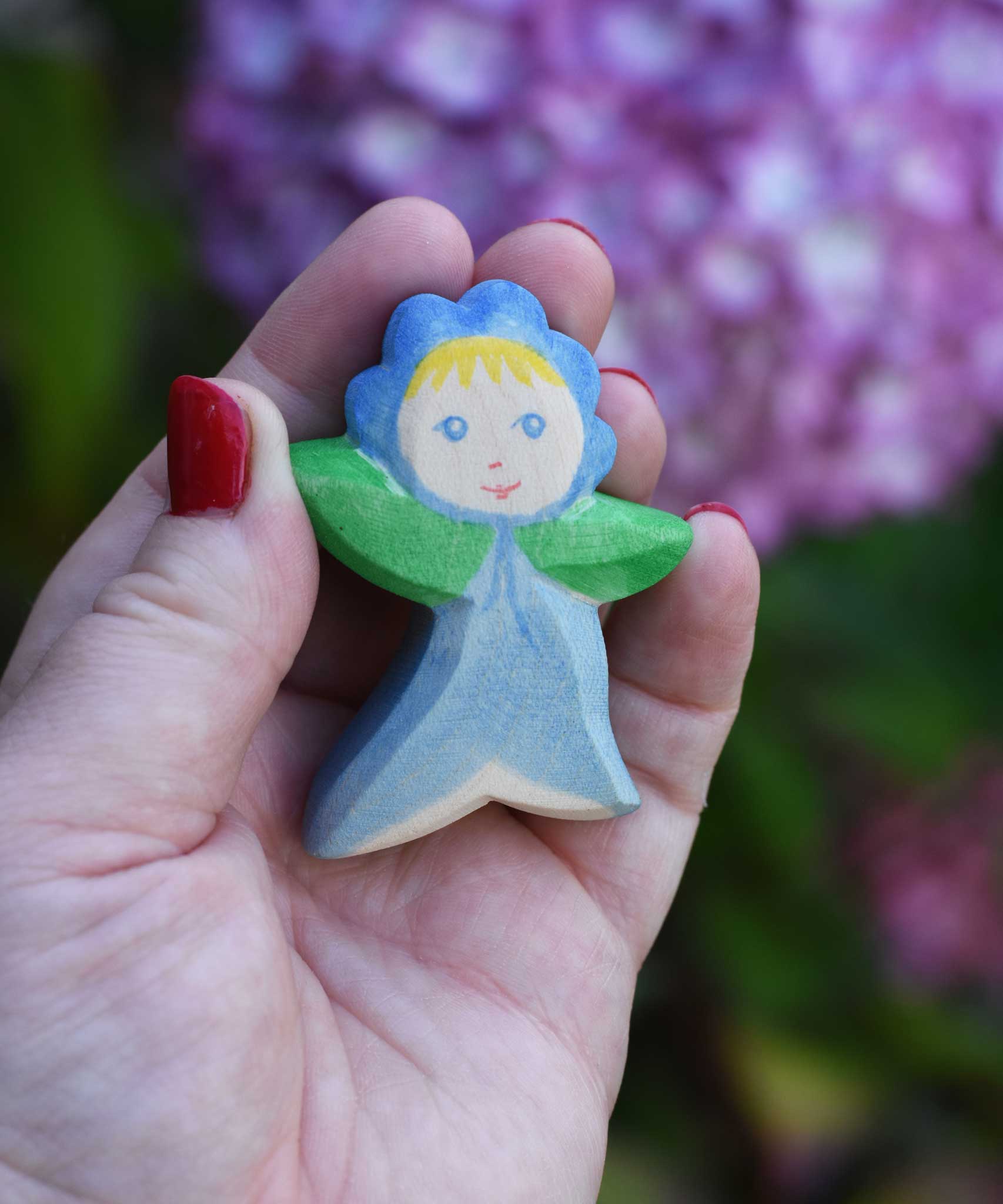 An Ostheimer hand made and hand painted wooden forget-me-not Flower Child figure toy held in an adult's hand to show scale. These flower child wooden figures are part of a wide range of expertly hand crafted wooden toys here at Babipur. 