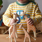 Ostheimer giraffe family handmade wooden toy figures. Child is holding a running and standing giraffe with a small with head low and small with head high giraffes on a wooden surface in front of them. 