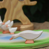 Ostheimer white goose with low neck wooden toy figure placed on an Ostheimer pond scene piece. Other wooden geese figures can be seen in the background along with the bird tree. 