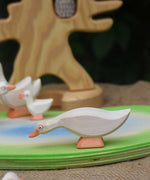 Ostheimer white goose with low neck wooden toy figure placed on an Ostheimer pond scene piece. Other wooden geese figures can be seen in the background along with the bird tree. 