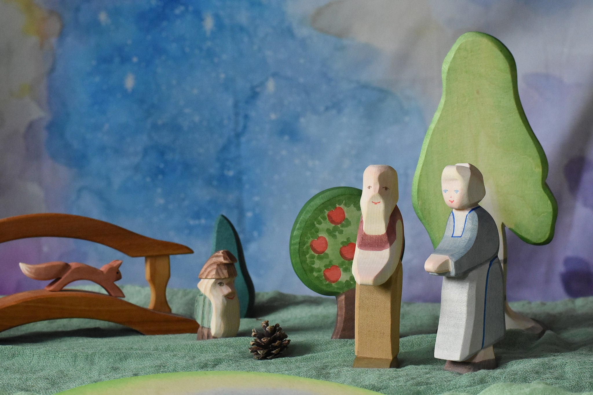 An Ostheimer grandmother and grandfather figure placed in a small world play scene. There is a green coloured decorative cloth covering the surface beneath them and an green bush, apple tree, large birch tree, fire tree dwarf and wooden bridge in the background.  These wooden figures are part of a wide range of expertly hand crafted wooden toys here at Babipur. 