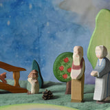 An Ostheimer grandmother and grandfather figure placed in a small world play scene. There is a green coloured decorative cloth covering the surface beneath them and an green bush, apple tree, large birch tree, fire tree dwarf and wooden bridge in the background.  These wooden figures are part of a wide range of expertly hand crafted wooden toys here at Babipur. 