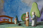 An Ostheimer grandmother and grandfather figure placed in a small world play scene. There is a green coloured decorative cloth covering the surface beneath them and an green bush, apple tree, large birch tree, fire tree dwarf and wooden bridge in the background.  These wooden figures are part of a wide range of expertly hand crafted wooden toys here at Babipur. 