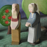 An Ostheimer grandmother and grandfather figure placed in a small world play scene. There is a green coloured decorative cloth covering the surface beneath them and an apple tree and large birch tree in the background.  These wooden figures are part of a wide range of expertly hand crafted wooden toys here at Babipur. 
