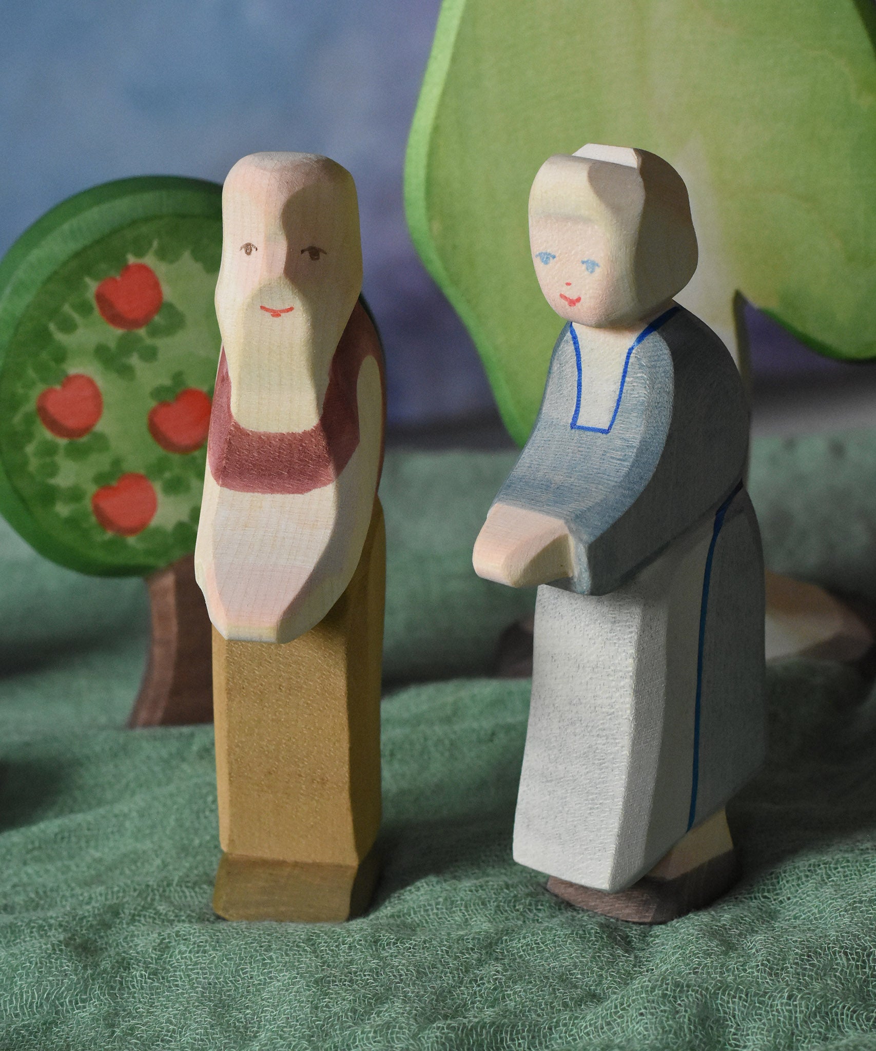 An Ostheimer grandmother and grandfather figure placed in a small world play scene. There is a green coloured decorative cloth covering the surface beneath them and an apple tree and large birch tree in the background.  These wooden figures are part of a wide range of expertly hand crafted wooden toys here at Babipur. 
