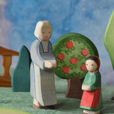 An Ostheimer grandmother figure and little red riding hood figure placed in a small world play scene. There is a green coloured decorative cloth covering the surface beneath them and an apple tree and green bush in the background.  These wooden figures are part of a wide range of expertly hand crafted wooden toys here at Babipur. 