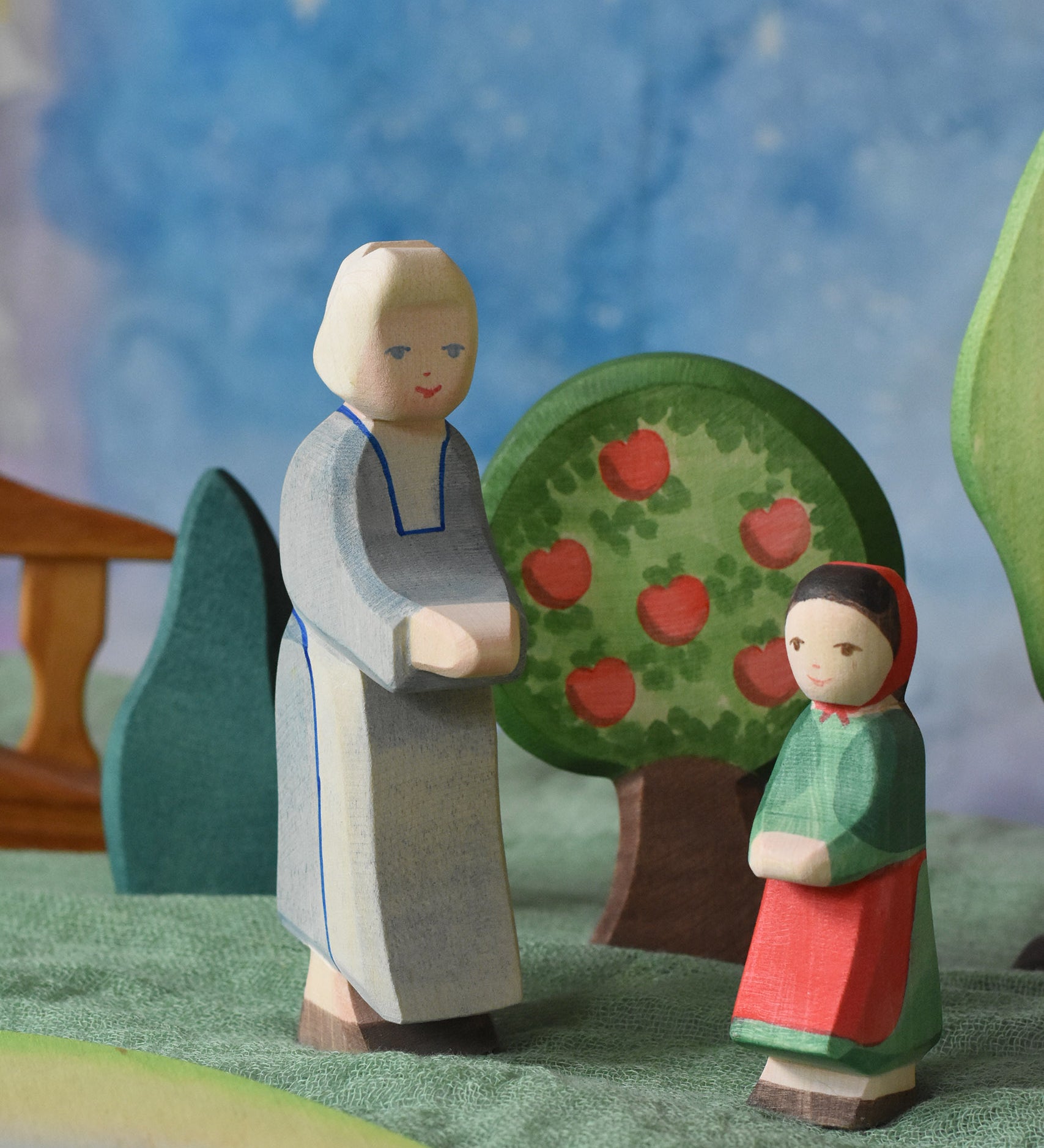An Ostheimer grandmother figure and little red riding hood figure placed in a small world play scene. There is a green coloured decorative cloth covering the surface beneath them and an apple tree and green bush in the background.  These wooden figures are part of a wide range of expertly hand crafted wooden toys here at Babipur. 