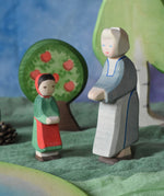 An Ostheimer grandmother figure placed next to a little red riding hood figure in a small world play scene. There is a green coloured decorative cloth covering the surface beneath them and an apple tree and large birch tree in the background.  These wooden figures are part of a wide range of expertly hand crafted wooden toys here at Babipur. 