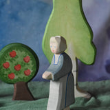 An Ostheimer grandmother figure placed in a small world play scene. There is a green coloured decorative cloth covering the surface beneath them and an apple tree and large birch tree in the background.  These wooden figures are part of a wide range of expertly hand crafted wooden toys here at Babipur. 
