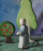An Ostheimer grandmother figure placed in a small world play scene. There is a green coloured decorative cloth covering the surface beneath them and an apple tree and large birch tree in the background.  These wooden figures are part of a wide range of expertly hand crafted wooden toys here at Babipur. 
