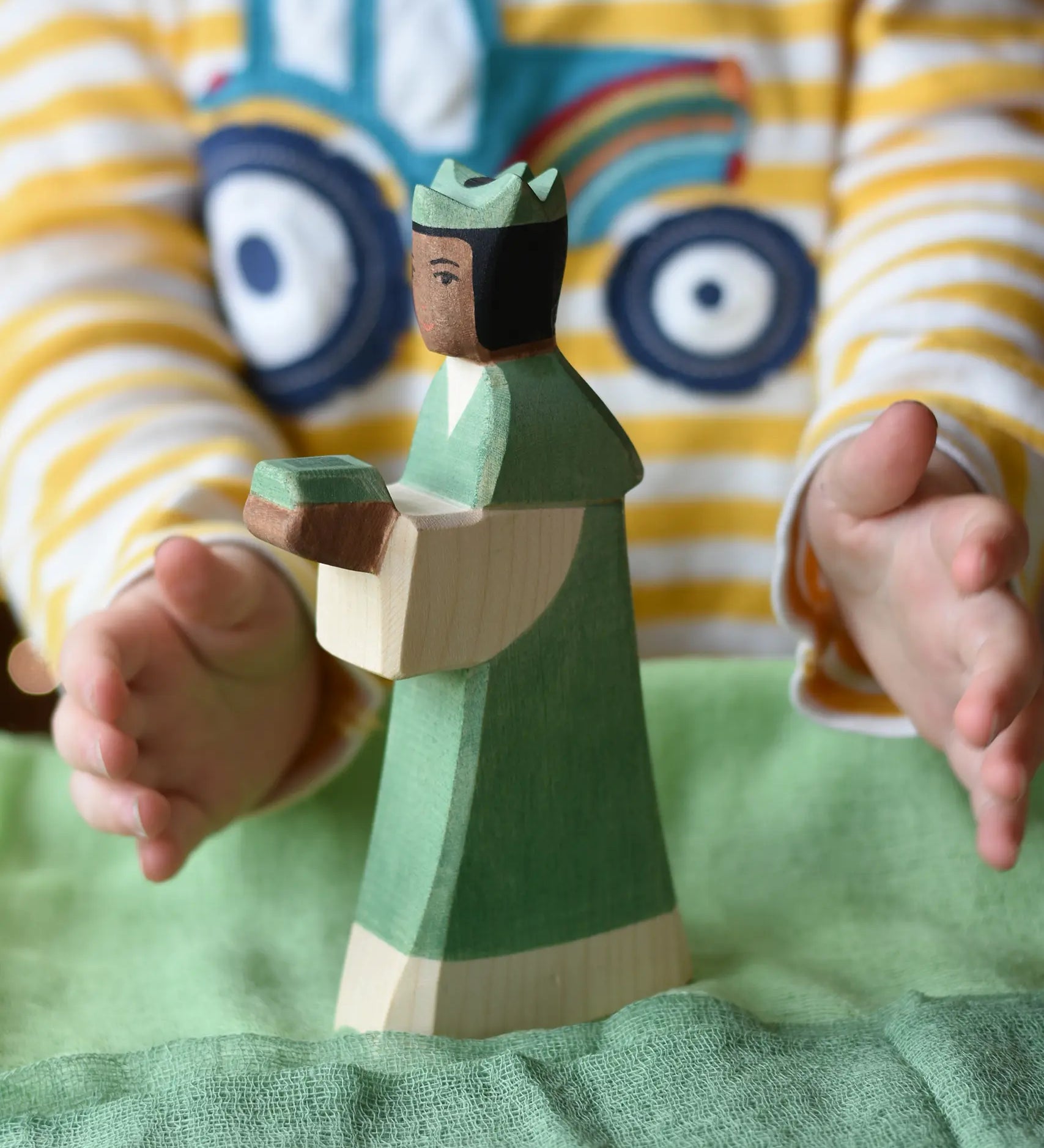 Ostheimer green king wooden toy figure placed on a cotton green play cloth with a child's hand going in to hold it. 