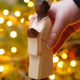 The Nativity Man figure by Ostheimer, a hand carved and hand painted man figure in simple cream robes and a hat, with dark brown skin and black hair. The figure toy is held in front of a Christmas tree.