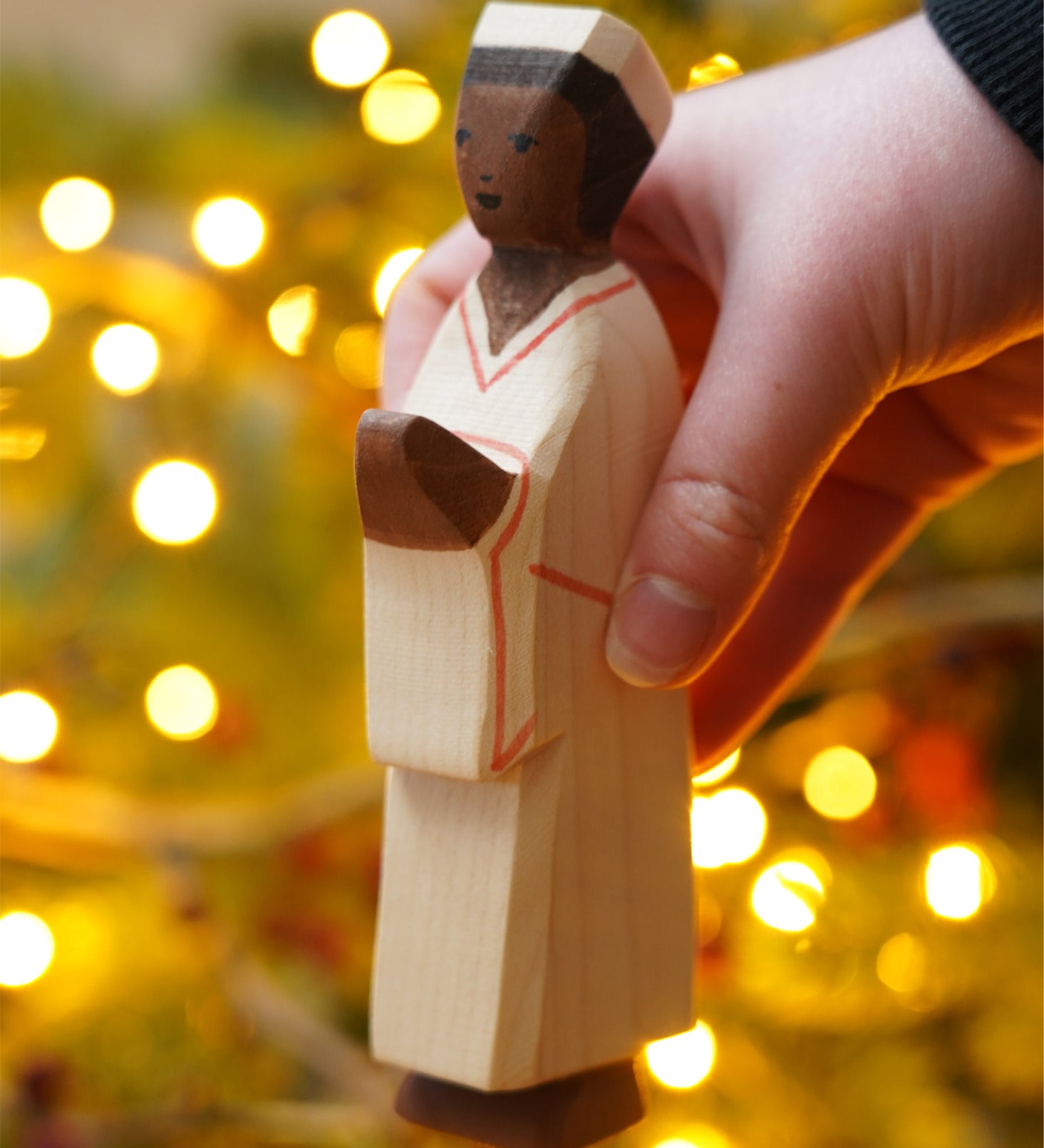 The Nativity Man figure by Ostheimer, a hand carved and hand painted man figure in simple cream robes and a hat, with dark brown skin and black hair. The figure toy is held in front of a Christmas tree.