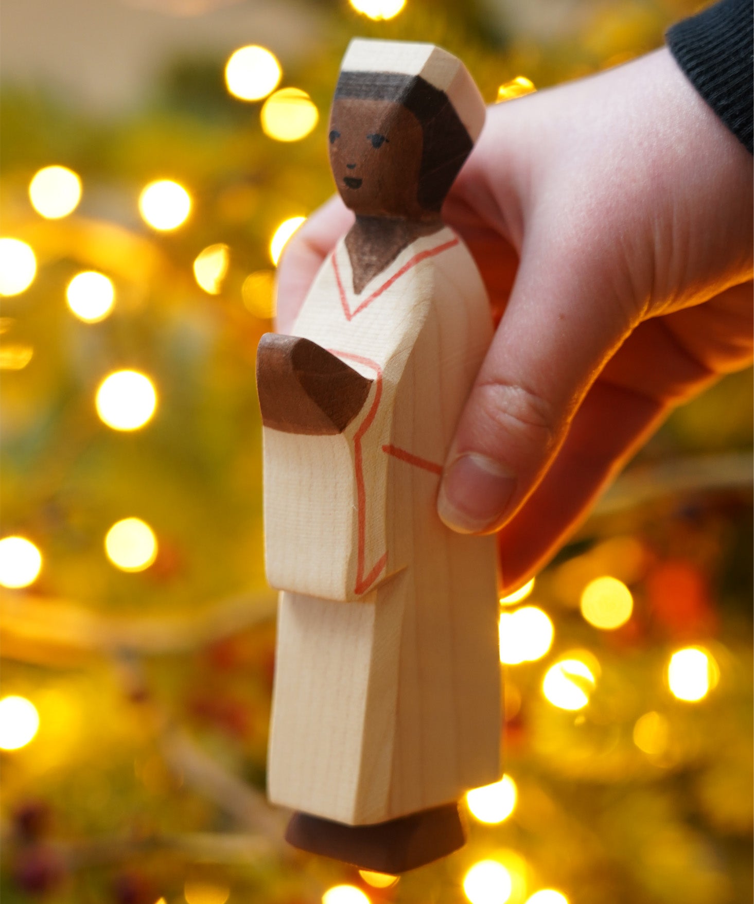 The Nativity Man figure by Ostheimer, a hand carved and hand painted man figure in simple cream robes and a hat, with dark brown skin and black hair. The figure toy is held in front of a Christmas tree.