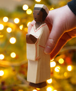 The Nativity Man figure by Ostheimer, a hand carved and hand painted man figure in simple cream robes and a hat, with dark brown skin and black hair. The figure toy is held in front of a Christmas tree.