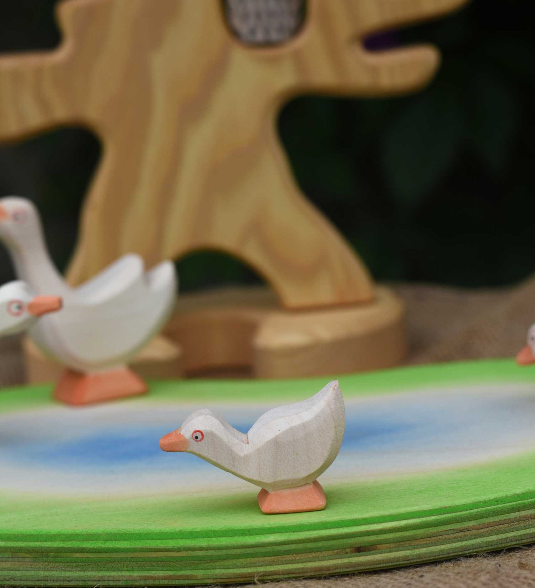 An Ostheimer gosling with it's head low placed on a pond scene piece. Other Ostheimer geese and a wooden bird tree can be seen in the background.