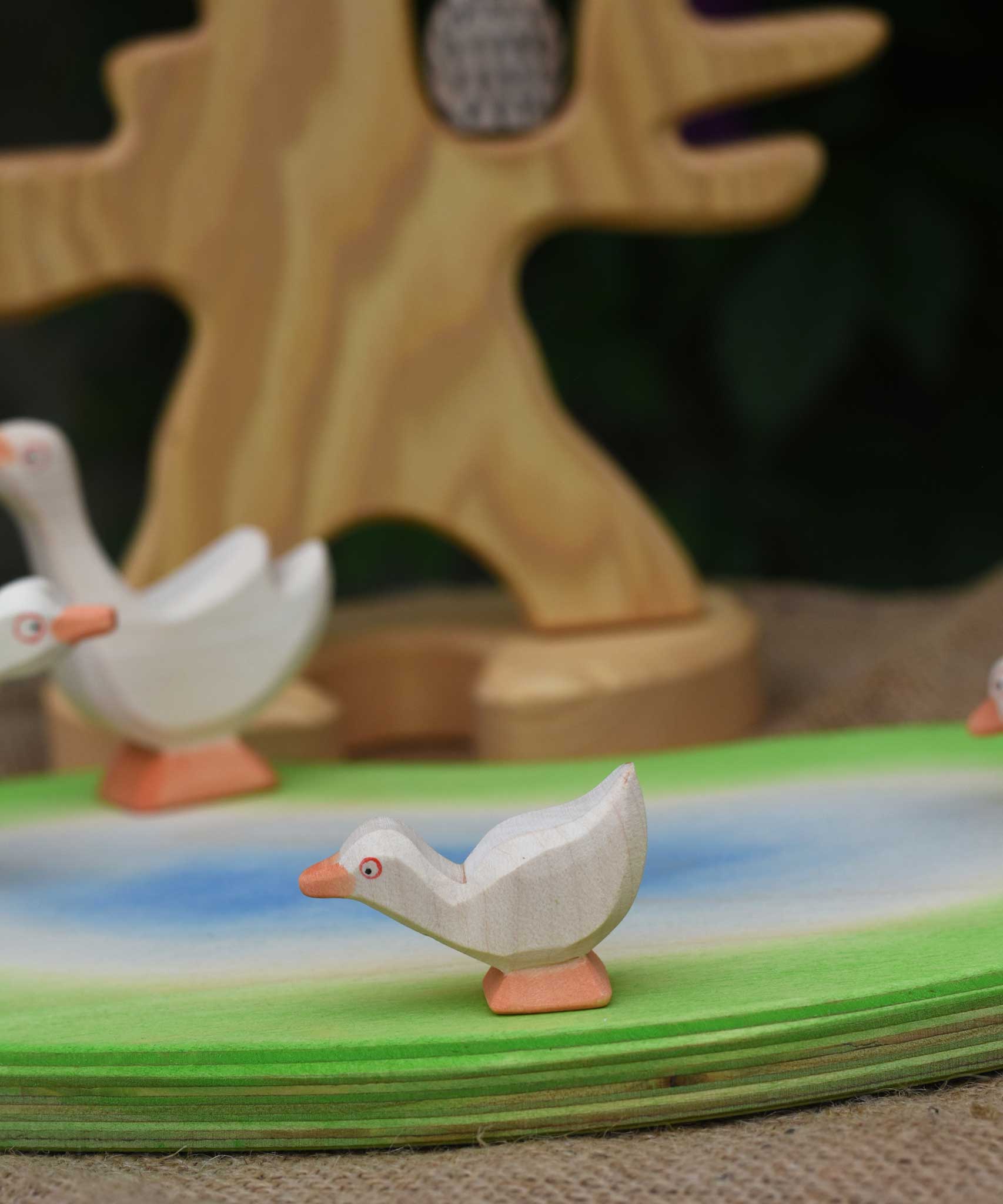 An Ostheimer gosling with it's head low placed on a pond scene piece. Other Ostheimer geese and a wooden bird tree can be seen in the background.