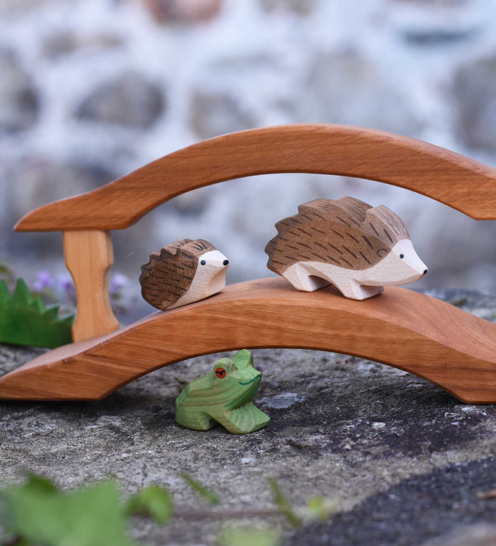Ostheimer hedgehogs, large and small placed on a wooden bridge with a green frog underneath. Check out our full range of expertly crafted wooden toy figures and scenery here at Babipur.