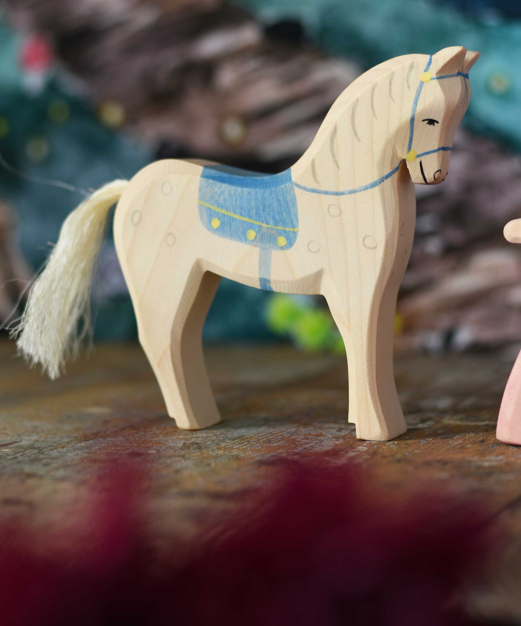 An Ostheimer white horse figure with blue coloured saddle placed on a wooden surface. These wooden figures are part of a wide range of expertly hand crafted wooden toys here at Babipur. 