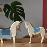 Two Ostheimer wooden white horse with blue saddle figures placed on a wooden surface with the coach house in the background.