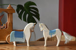 Two Ostheimer wooden white horse with blue saddle figures placed on a wooden surface with the coach house in the background.