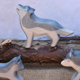 An Ostheimer Howling Wolf wooden toy figure shown in a small world scene set up with other Ostheimer wolves. The wolf is standing on a log branch. These figures are part of a vast range of expertly crafted wooden toy figures and scenery here at Babipur.