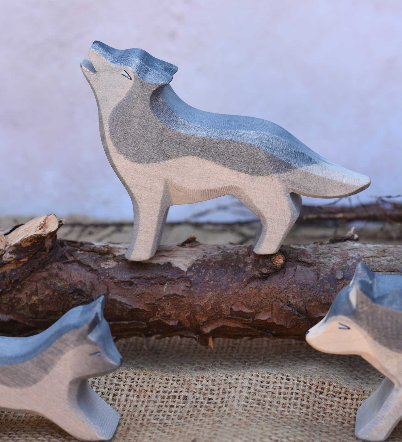 An Ostheimer Howling Wolf wooden toy figure shown in a small world scene set up with other Ostheimer wolves. The wolf is standing on a log branch. These figures are part of a vast range of expertly crafted wooden toy figures and scenery here at Babipur.